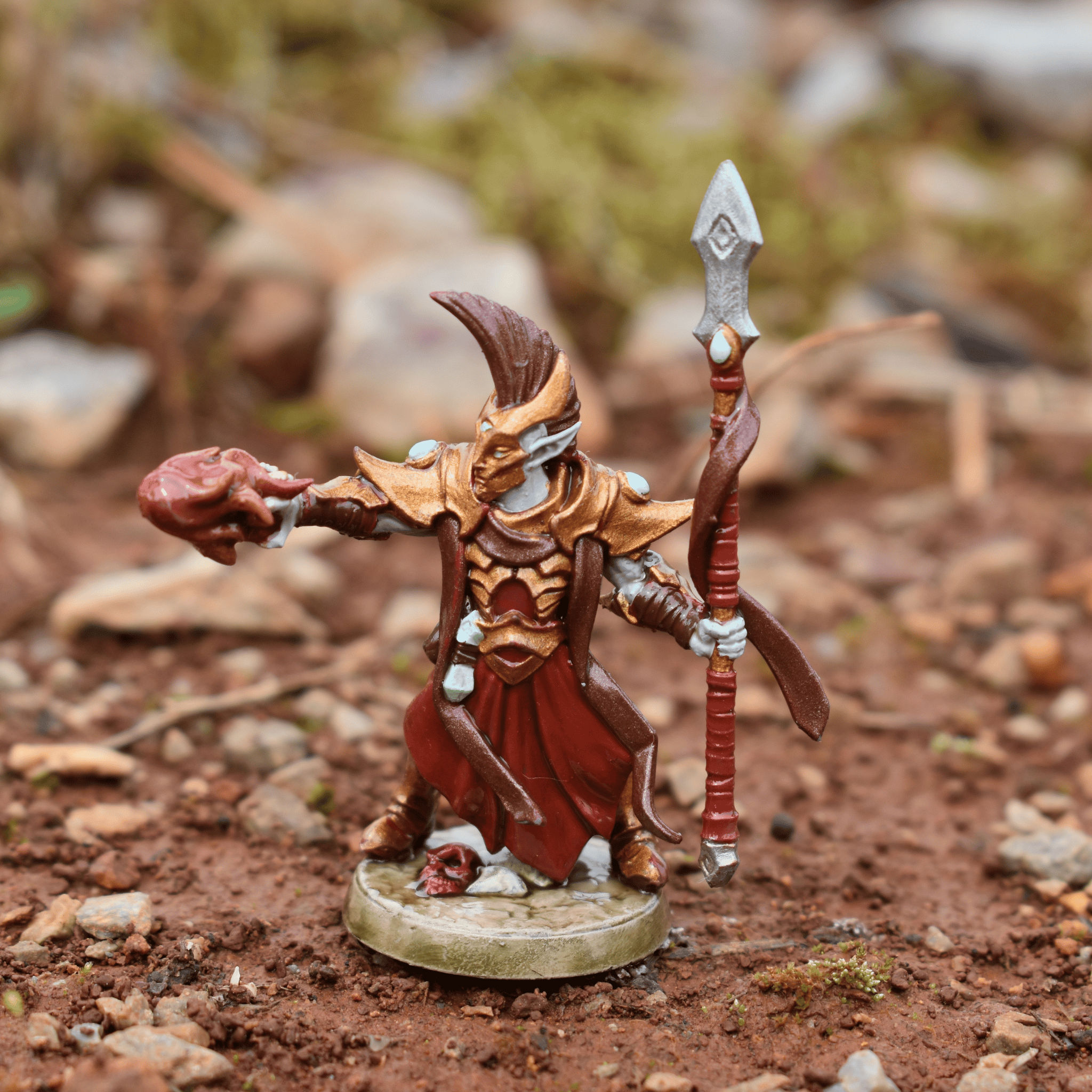 Painted miniature of a red and gold armored mage holding a heart and a staff, displayed on rocky terrain.