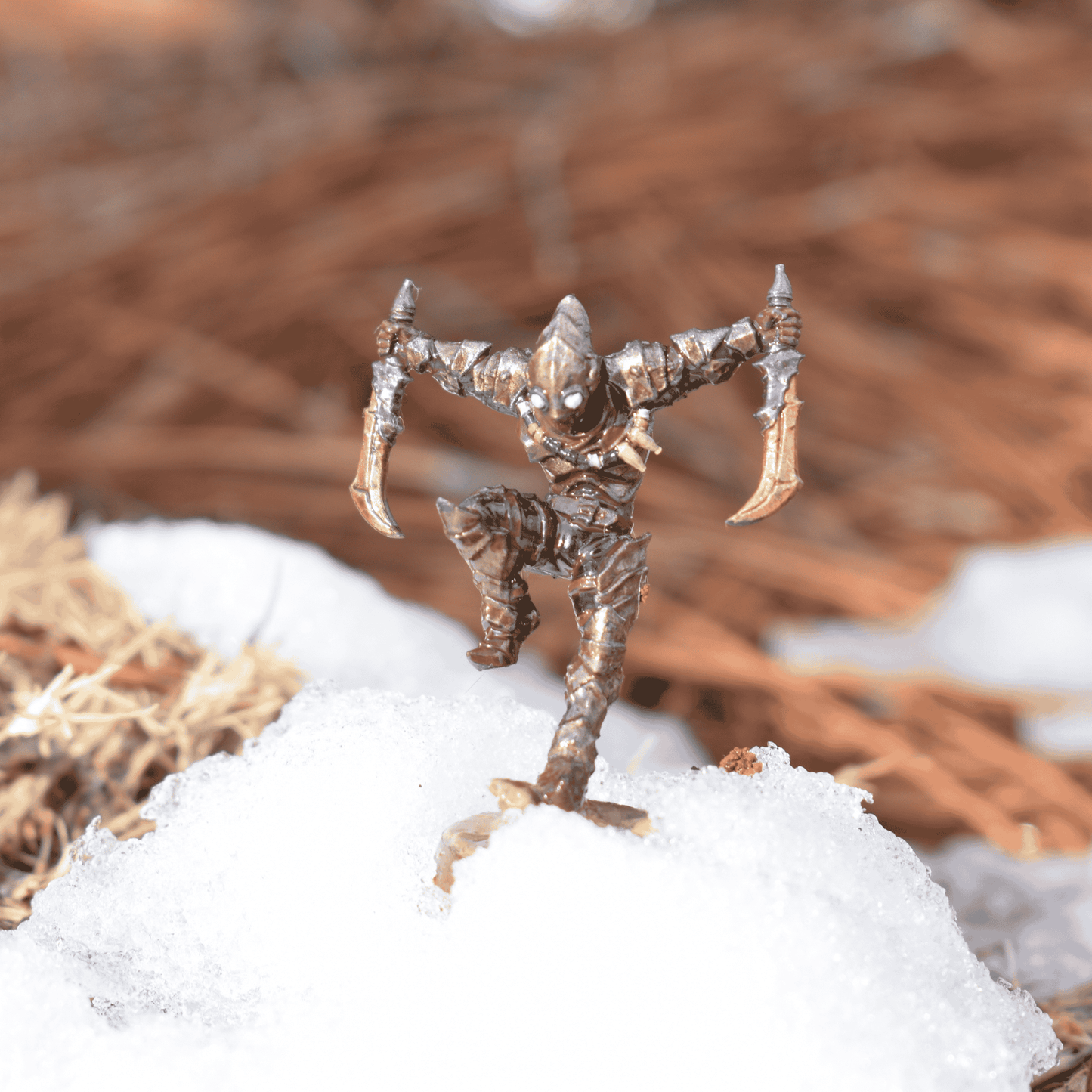 Fantasy assassin miniature standing in a patch of snow, dual blades raised, with bronze armor and glowing green eyes.