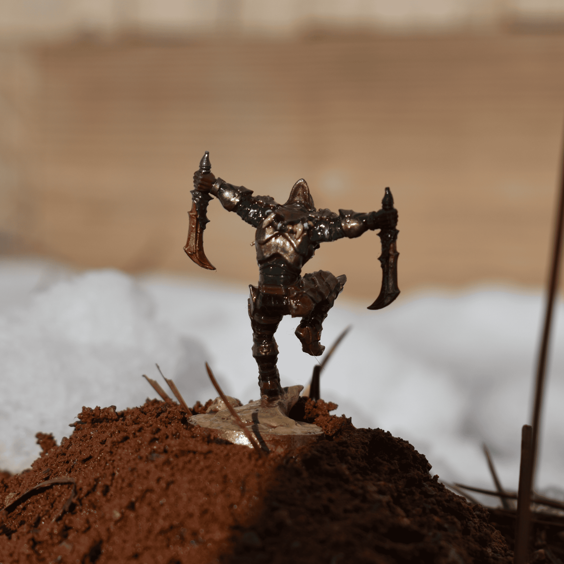 Rear view of an armored assassin miniature in a striking stance, displayed on red earth and flanked by melting snow.