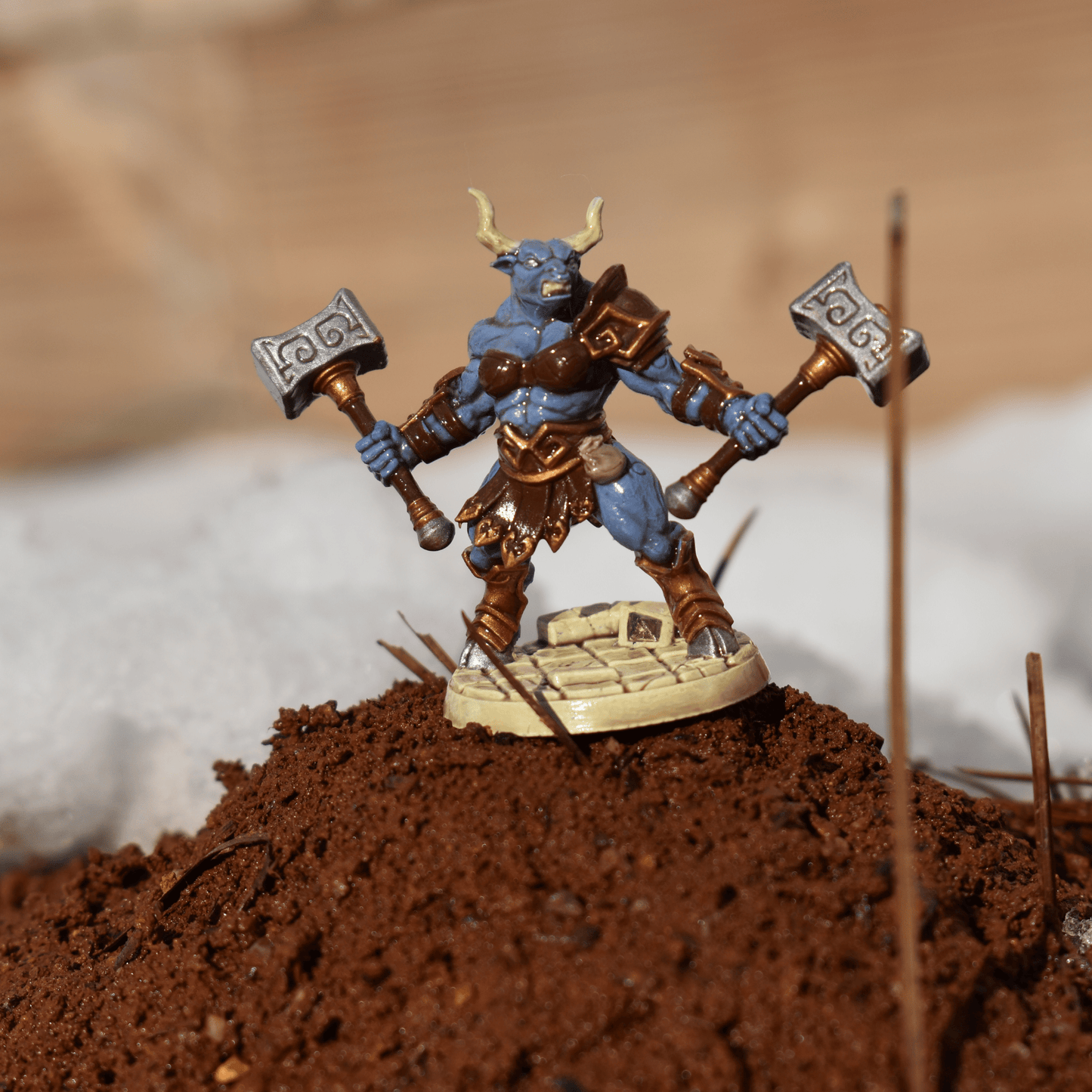 Fantasy Minotaur miniature posed on a raised patch of dirt, wearing brown armor and wielding dual hammers, with snowy background elements.