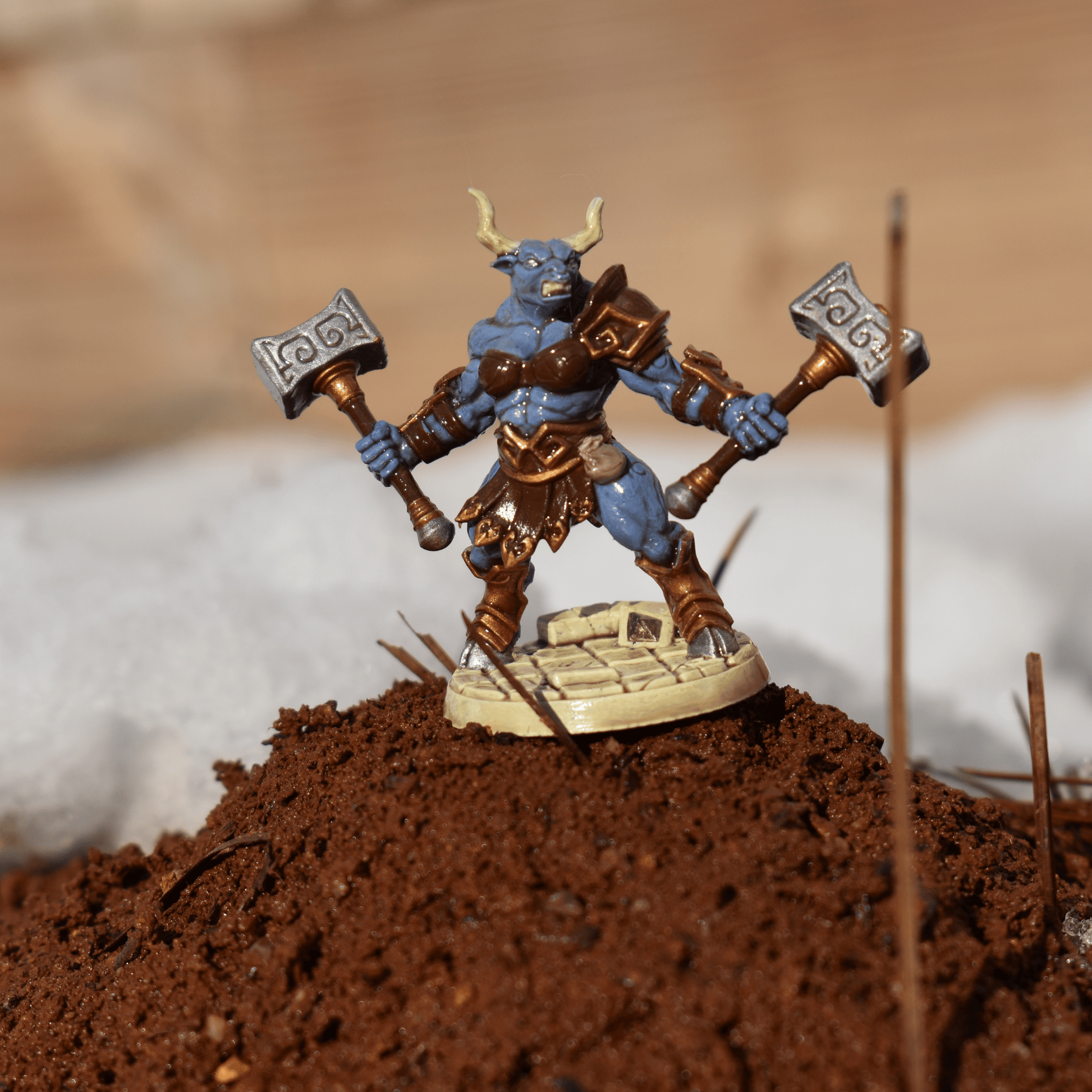 Fantasy Minotaur miniature posed on a raised patch of dirt, wearing brown armor and wielding dual hammers, with snowy background elements.