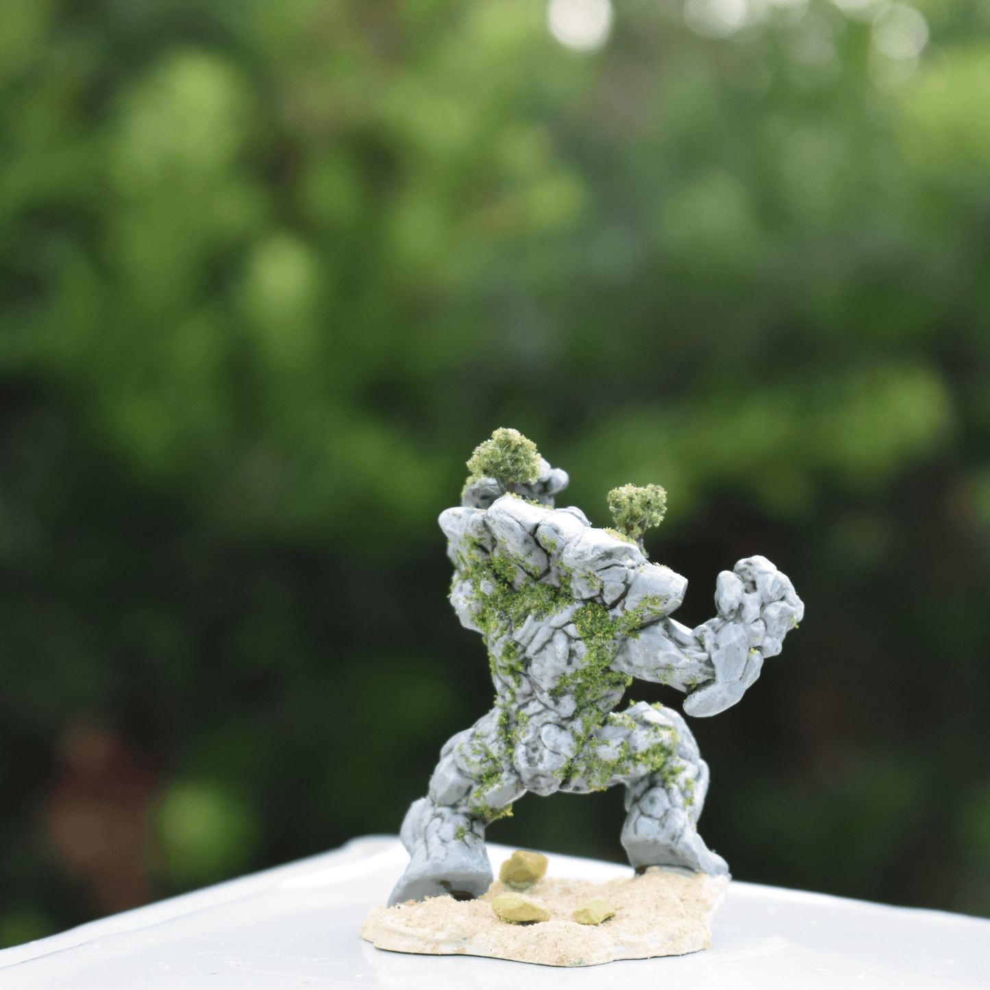 Back view of a hand-painted Earth Elemental miniature with moss and tree detailing, standing on a textured sand base against a green background.