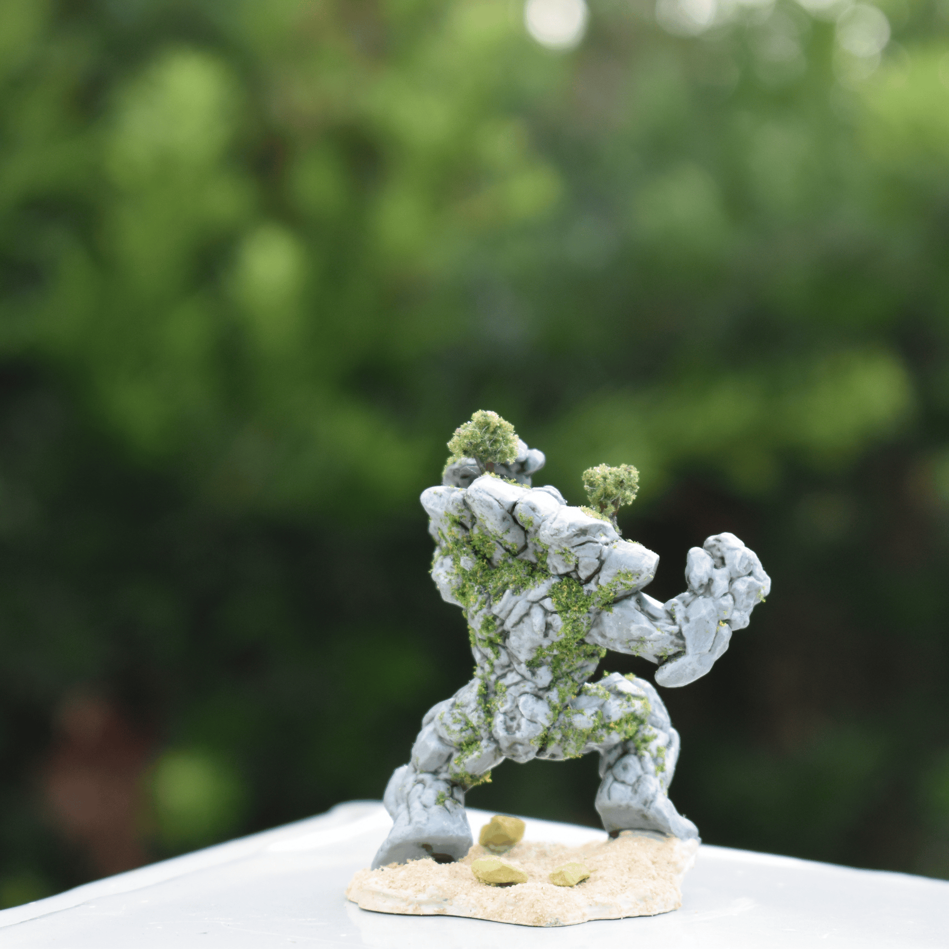 Back view of a hand-painted Earth Elemental miniature with moss and tree detailing, standing on a textured sand base against a green background.