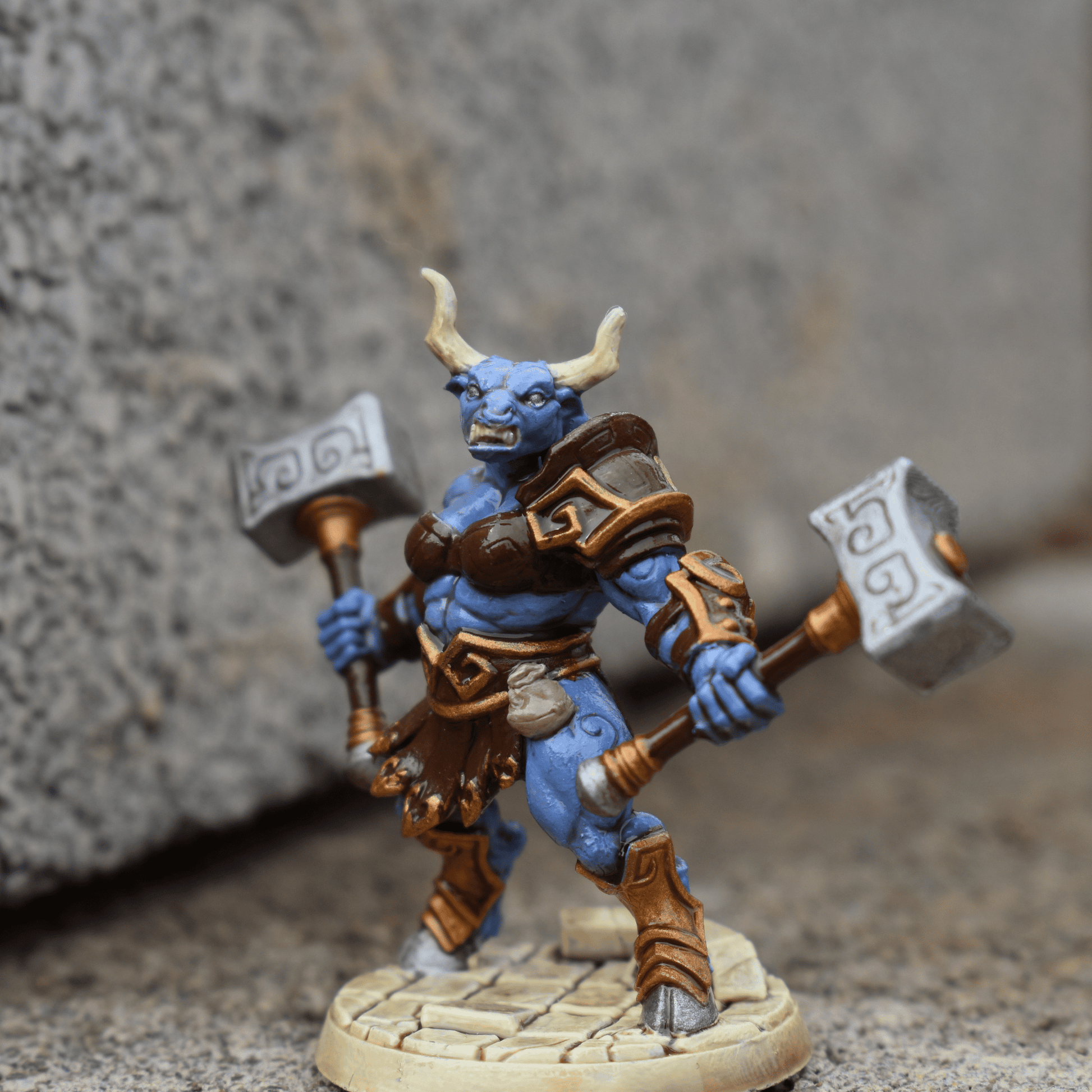 Hand-painted Minotaur warrior miniature with blue skin and gold-brown armor, holding twin rune-covered hammers, posed in front of a stone wall.
